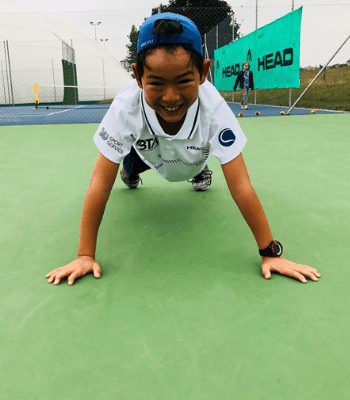 Petit enfant faisant la planche et souriant sur un court de tennis en extérieur, avec le t-shirt BTA