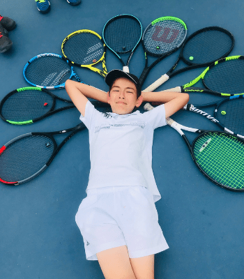Petit garçon couché sur le sol d'un court de tennis, plusieurs raquettes entourant sa tête